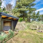 Ein kleines Holzhaus mit Sitzbank und Feuerstelle im Gras unter Bäumen.