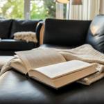 Ein offenes Buch liegt auf einem schwarzen Ledersofa mit einer hellen Decke.