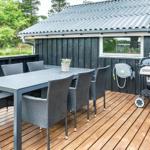 Terrasse mit Tisch, Stühlen und Grill. Hintergrund: dunkler Holzaufbau und Grün.