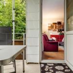 Terrasse mit Tisch und Stuhl. Durch Glastür ist Wohnzimmer mit rotem Sofa sichtbar.