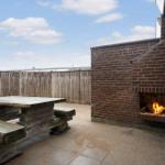Außenbereich mit Holztisch, Grill und Steinplatten. Hintergrund: Holzzaun und blauer Himmel.