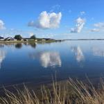 Stille Wasserfläche spiegelt Himmel und Häuser wider. Windräder sind im Hintergrund sichtbar.
