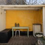 Yellow outdoor area with table, stool, and plants under roof.