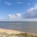 Strand mit Sand, Gras und Meer unter blauem Himmel mit weißen Wolken.