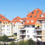 Mehrfamilienhaus mit rotem Dach und Balkonen unter blauem Himmel.
