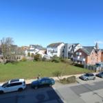 Ansicht über eine grüne Wiese mit geparkten Autos und Wohnhäusern unter blauem Himmel.