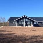 Moderne Villa mit großem Glasbereich und dunklem Holzverkleidung. Vorhof mit Erde und Bäumen im Hintergrund.