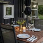 Gedeckter Holztisch mit Stühlen auf Terrasse vor dunklem Haus mit Fenstern.