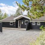 Black wooden house with roof and windows. Front yard with gravel and trees.