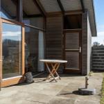 Terrace with table and chair in front of wooden house with glass facade and entrance door.