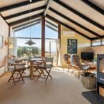 Living and dining area with wooden table, chairs, and wood stove. Large windows with view of landscape.