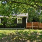 Schwarzes Holzhaus mit weißen Fensterrahmen, umgeben von Bäumen und Gras.