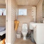 Badezimmer mit Toilette, Waschbecken und Fenster. Holzwände und -boden. Bett im Hintergrund.