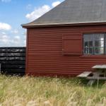 Rotes Holzhaus mit Picknicktisch und Stuhl im Gras. Fenster mit Glasteilen.