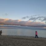 Zwei Personen und ein Hund spazieren am Strand entlang bei Sonnenuntergang.