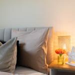 Bettkopf mit Kissen und Nachttisch mit Lampe und Blumen.