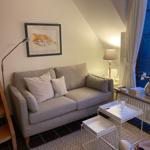 Ein Wohnbereich mit Sofa, Tischen und Fenster. Wandbild und Stehlampe sind sichtbar.