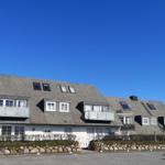 Ein zweistöckiges Haus mit grauem Dach und weißen Fenstern. Vor dem Haus befindet sich ein Steinwall.