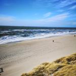 Strand mit Wellen, Sand und Dünenvegetation unter blauem Himmel.