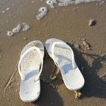 Weiße Flip-Flops liegen am sandigen Strand mit Seegras und Schaum.