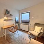 Ein Arbeitszimmer mit Schreibtisch, Stuhl und Sessel. Fenster, Wandregale und Teppich sind sichtbar.