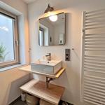 Badezimmer mit Waschbecken, Spiegel und Fenster. Links steht ein Pflanzenkorb.