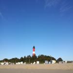 Rote Leuchtturm mit weißen Streifen auf sandigem Strand unter blauem Himmel.
