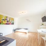 Zimmer mit Bett, Esstisch und Wandbild. Holzboden und Fenster.