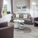 Wohnzimmer mit Sofa, zwei Sesseln und Wohnwand. Fenster mit Vorhängen und Wandbild.