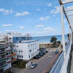 Blick von Balkon auf Strand, Parkplatz und Meer unter blauem Himmel mit weißen Wolken.