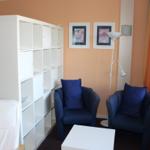 Zimmer mit zwei blauen Sesseln, weißem Tisch und Regal. Fenster mit orangefarbenen Vorhängen.