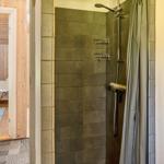 Dusche mit Duschbrause und Regal. Tür führt in Schlafzimmer mit Bett und Fenster.