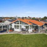 Modernes Haus mit großem Holzterrasse und rotem Dach. Grünfläche und Gartenbereich.