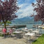 Terrasse mit Tischen und Stühlen am See mit Booten und Bergen im Hintergrund.