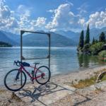 Ein rotes Fahrrad steht an einem Seeufer mit Bergen im Hintergrund.