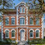 Rotes Backsteinhaus mit weißen Fensterrahmen und Eingangstür. Umgeben von Bäumen und Grünflächen.