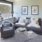 Ein Wohnzimmer mit grauem Sofa, Sessel und Glas-Tischen. Blick auf eine Terrasse mit Berglandschaft.