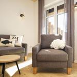 Ein Wohnzimmer mit grauem Sofa und Sessel, Holztisch und Fenster mit Vorhängen.