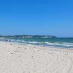 Strand mit weißen Sand, Wellen und klarem Himmel. Zwei Personen spazieren entlang des Wassers.