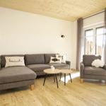 Wohnzimmer mit grauem Sofa, Sessel und Holzboden. An der Wand hängen zwei Lampen.