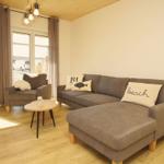 Wohnzimmer mit grauem Sofa, kleinem Tisch und Fenstern mit Vorhängen.