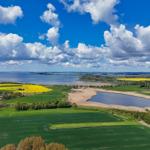 Weitläufige Landschaft mit Feldern, einem See und dem Meer unter blauem Himmel mit weißen Wolken.
