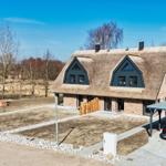Modernes Haus mit Strohdach und Carport. Freier Platz und Bäume im Hintergrund.