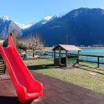 Roter Rutschbahn und Spielhütte am See mit Bergen im Hintergrund.