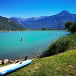 Ein Stand-Up-Paddleboard liegt am Ufer eines türkisfarbenen Sees mit Bergen im Hintergrund.