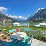 Terrasse mit Tisch und Stühlen am See mit Bergen im Hintergrund.