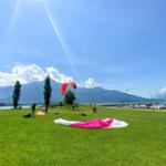 Grünes Feld am See mit Paragliders und Bergen im Hintergrund.