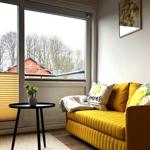 Gelber Sofa mit Kissen, kleiner Tisch und Fensterblick auf Bäume.