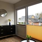 Schwarzer Schrank mit goldfarbenen Griffen und runder Spiegel an der Wand. Fenster mit Jalousie und Blick auf Dächer.
