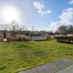 Grüner Rasenbereich mit Weg, Hecken und Bäumen unter blauem Himmel.